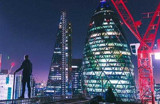 两个人站在楼顶上俯瞰伦敦的夜景，凝望着伦敦“小黄瓜”