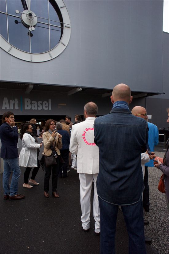 　　2016年瑞士巴塞尔艺博会入口，身着白色西装的参观者后背印有 “瑞士艺术巴塞尔（switzerland art basel）”字样。图片来源：马丽亚