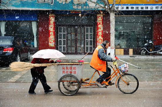 2013年2月18日，山西永济，纷飞的雪花中，一位64岁的环卫工人带着刚看完病的盲老伴回家，他边走边不忘捡拾地上的垃圾。