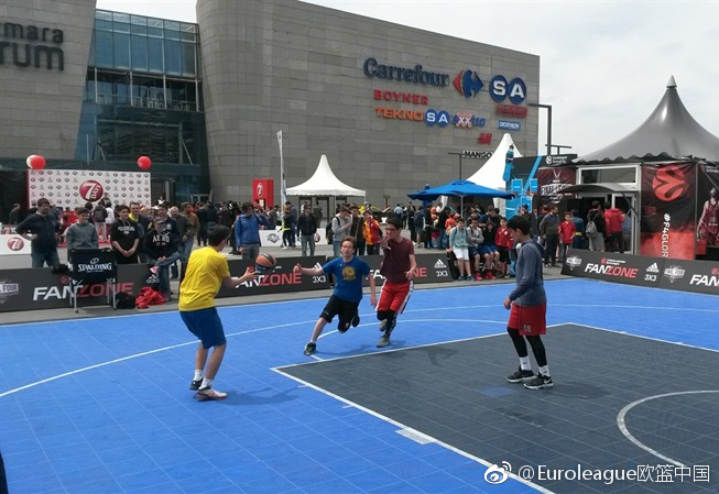 一群少年在球迷专区的迷你篮球场上腾空跃起，橘色皮球在空中划过弧线，背景是巨大的欧冠联赛标志，青春的活力和梦想在此刻定格。