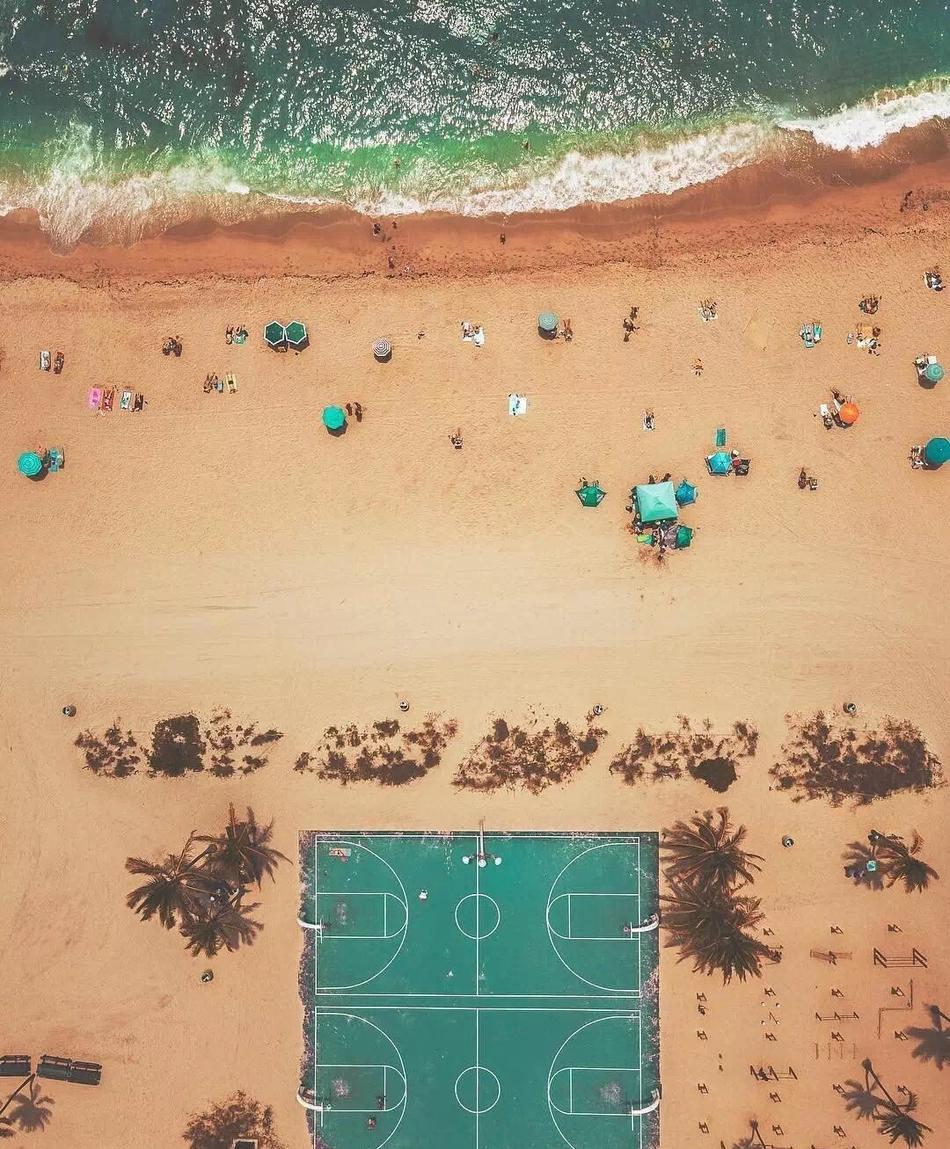 黄昏时分，几名少年在露天篮球场上打球，暖黄的灯光将他们包围，场边的绿树和远处的高楼剪影构成了宁静而充满活力的生活画卷。