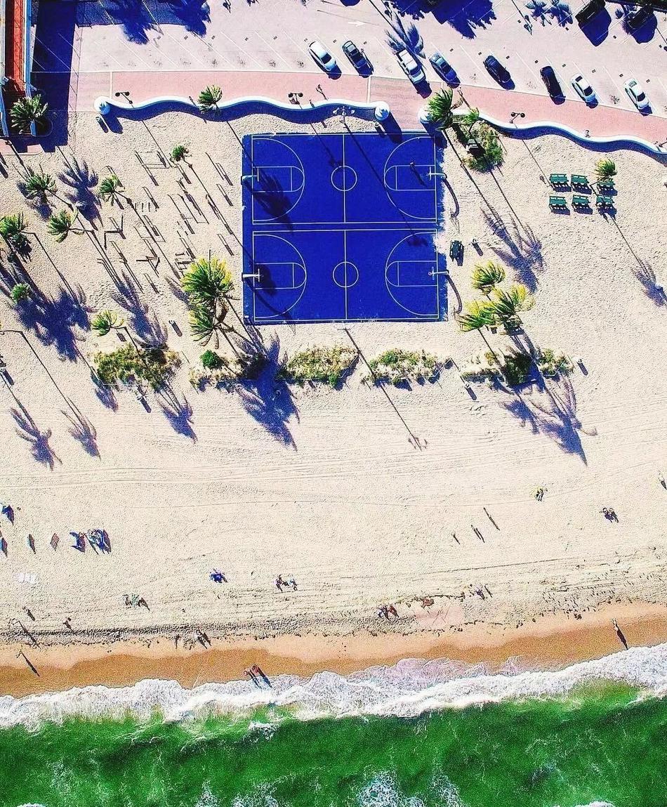 夕阳余晖下，从高处俯瞰篮球场，暖色调的灯光与蓝色地面形成鲜明对比，球员的影子被拉长，如同舞台上的舞者。