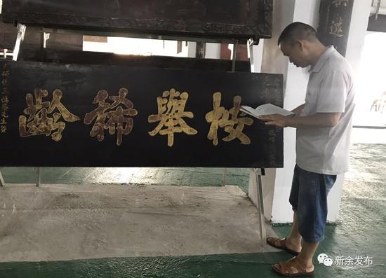 图为罗志鸿边讲解匾额，边查阅书籍。