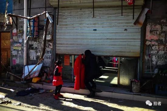 　　▲2017年11月22日，大兴区新建村，一位女士一手提着一件崭新的红色大衣，一边躬身拉上卷帘门，孩子在一旁等待离开。摄影/新京报记者彭子洋