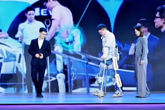 2025年保姆机器人大会现场，因意外事故导致下肢瘫痪的林寒（中）穿戴布法罗为其定制的外骨骼机器人展示无须借助外力实现行走。图据布法罗机器人科技。