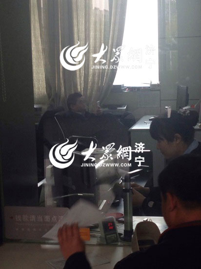 濟寧鄒城工商銀行的大廳內，柜員正在辦理業務，后面一位工作人員擺出腳踩辦公桌的姿勢，正在擺玩手機。