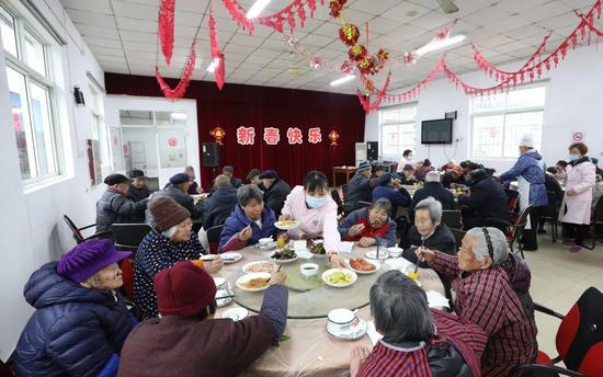 廊下镇敬老院的老人们围坐一起享用年夜饭，护工们化身“服务员”将菜传到饭桌上 。
