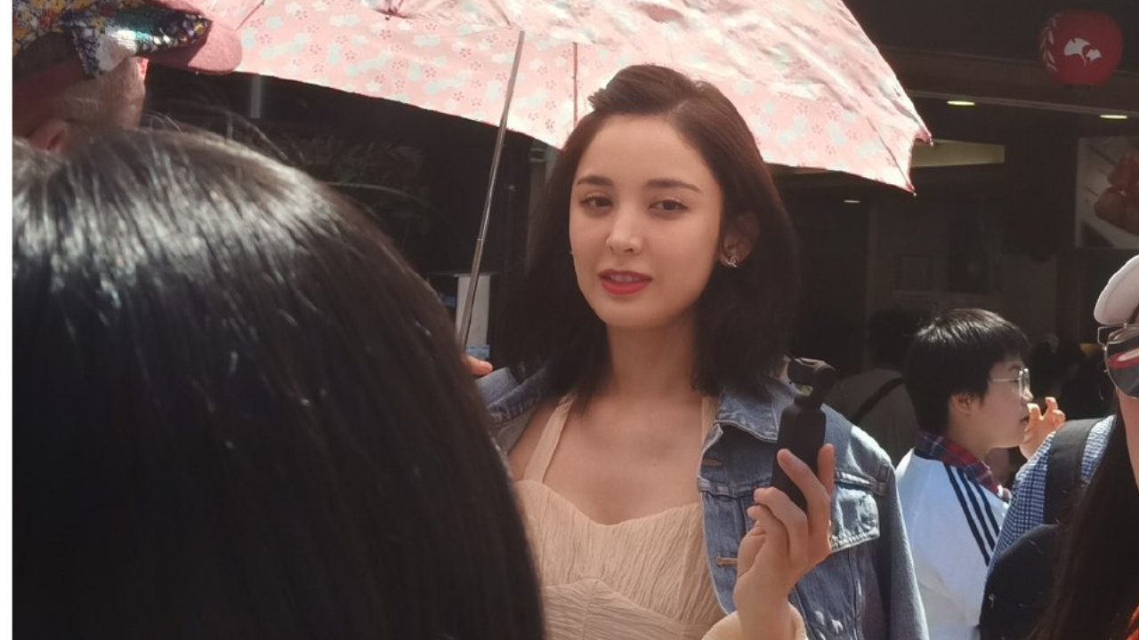 娜扎撑着粉色的遮阳伞，肩上披着牛仔褂穿一件白色吊带裙，皮肤白皙