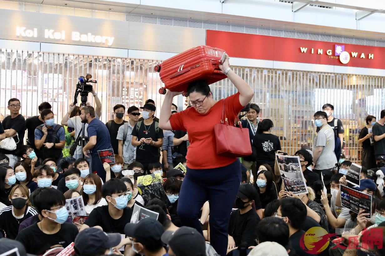 正扛着行李箱穿过非法示威者的女士，以及身后落闸停业的商铺。（图自文汇网）