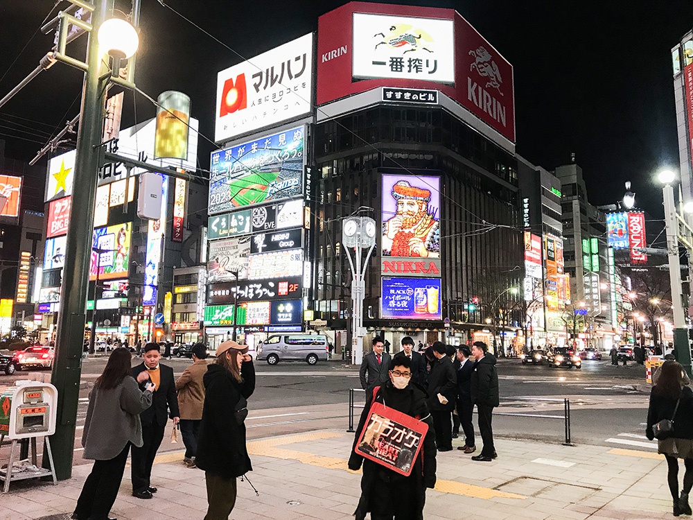 3月15日，日本北海道札幌薄野，夜晚的街头。画面正中戴着口罩的男子身上挂着一个广告牌，上面写着“卡拉OK”。日本之前部分群聚感染就是因卡拉OK引爆，因此现在其经济也较为萧条。画面后方穿西装的几名男子，则是像往常一样刚刚下班的上班族。北海道政府2月28日宣布进入疫情的紧急状态，于3月19日结束紧急状态。（摄影：李若涵）