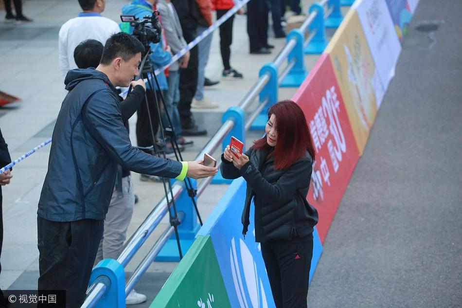 冬日娜身披央视外套,微笑着拿出手机,杜锋则弯下腰凑近屏幕,两人在喧闹的马拉松赛场边专注地扫描二维码,背景是模糊的人群和赛事横幅,画面充满了人情味。