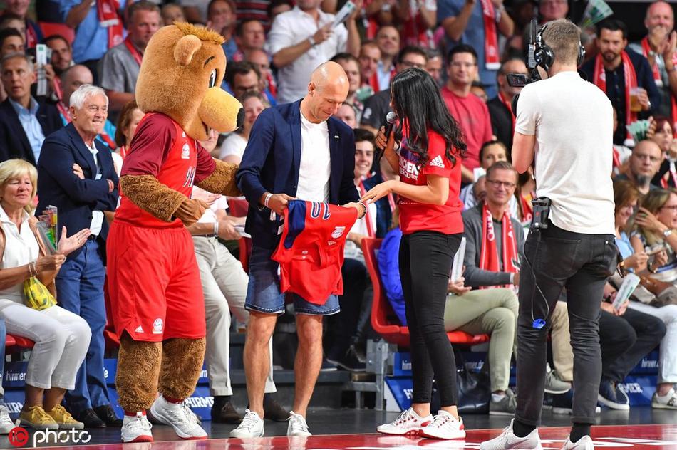 罗本面带温和笑容，手持拜仁篮球定制球衣，身旁的爱子略显腼腆地靠在父亲身前，背景是热闹的篮球馆看台，记录下这难忘瞬间。