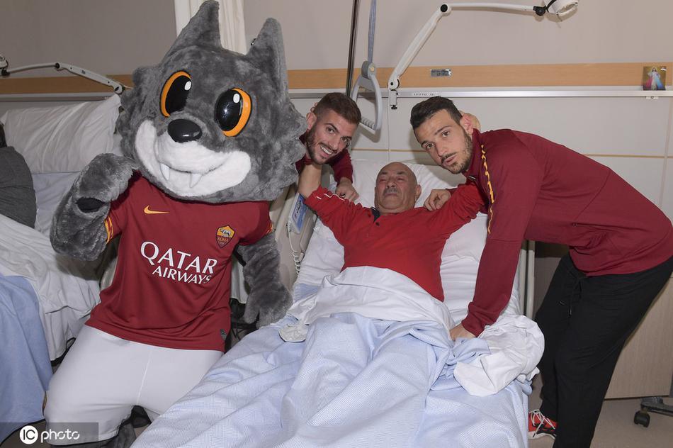 罗马队吉祥物这只毛茸茸的大灰狼正与一位老年患者亲切击掌，滑稽可爱的动作逗得老人开怀大笑，病房里洋溢着温馨与喜悦。