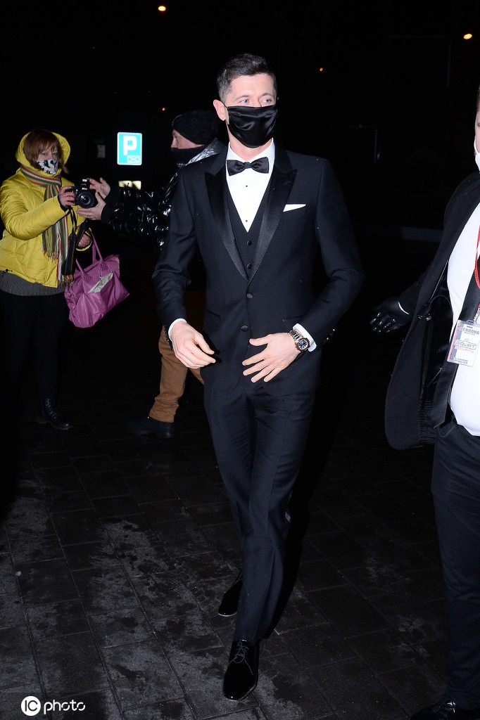 在闪光灯的照耀下，安娜侧身而立，精致的妆容和耳饰熠熠生辉，她低头整理裙摆，莱万则体贴地站在一旁，身形形成一个保护者的姿态