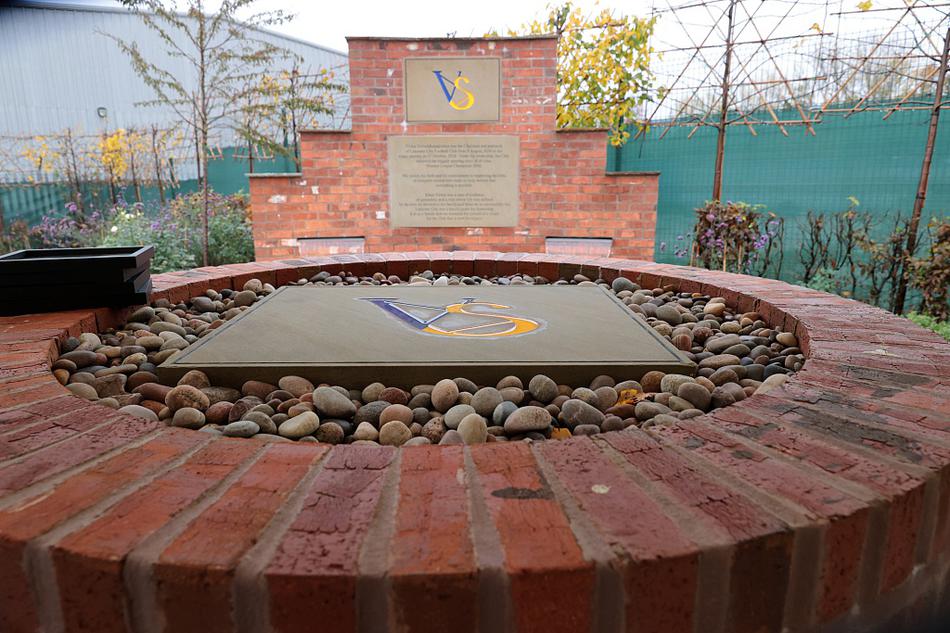 杰米·瓦尔迪神情凝重地站在维猜纪念花园，他身着深色西装，在鲜花和绿植的映衬下，目光中饱含着对昔日领路人的无限追思与敬意。