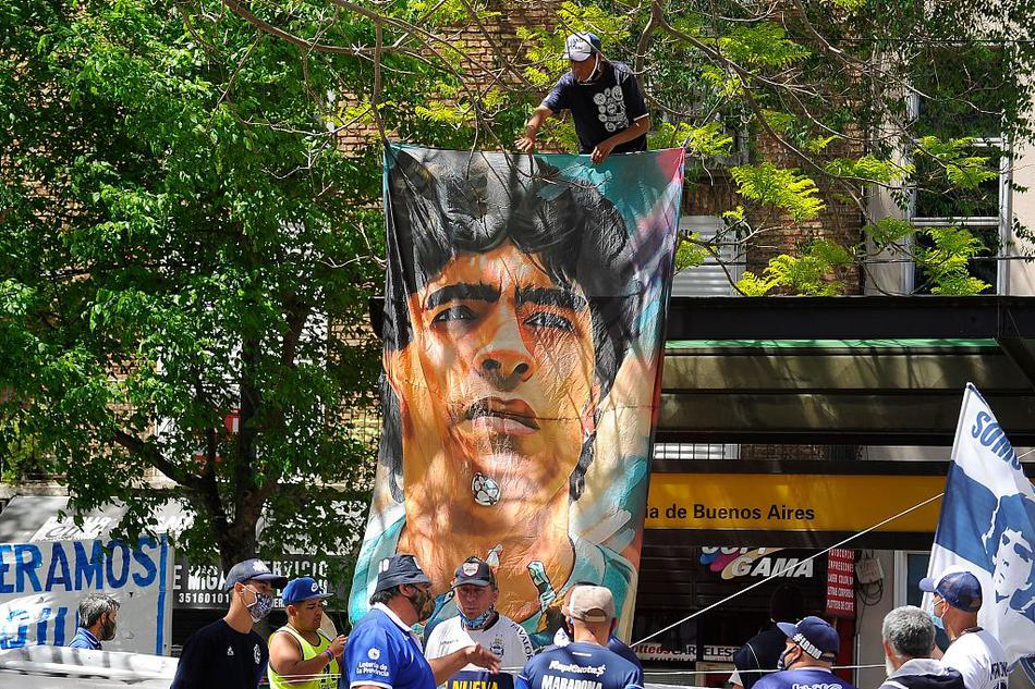 夜色中，两位球迷在医院外的铁栅栏前伫立，他们手持印有马拉多纳经典庆祝动作的旗帜，远远眺望着病房窗口那盏象征希望的灯光