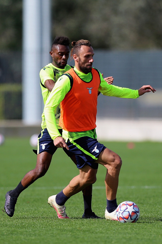 训练课尾声，一名拉齐奥球员坐在草地上进行拉伸，汗水浸湿了他的球衣。远处，教练组正在白板前讨论着明天的战术，这幅画面生动诠释了大战将至，全队上下众志成城的紧迫感。