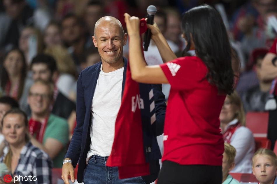 罗本双手展开刚刚获赠的拜仁篮球定制球衣，脸上洋溢着灿烂笑容，儿子在他身旁仰头观看，父子共享这份来自俱乐部的荣耀时刻。