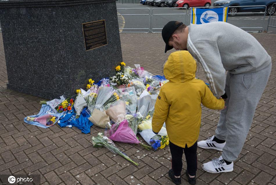 年轻的球迷带着孩子，蹲在雕像前仔细整理球迷留言卡，孩子的小手轻轻触碰雕像的球鞋，仿佛在通过这种方式，触摸那个从未见过面的偶像的余温。