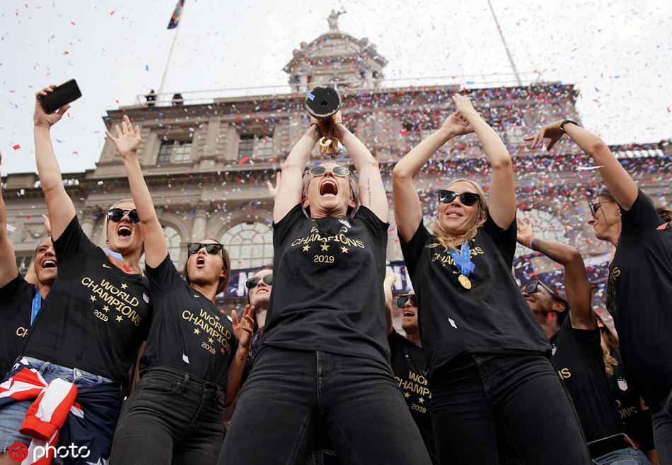 美国女足球星亚历克斯·摩根在花车上开怀大笑，她一手扶着栏杆，一手向人群比心，脸上洋溢着幸福的笑容，胸前挂着的金牌在阳光下闪闪发光。