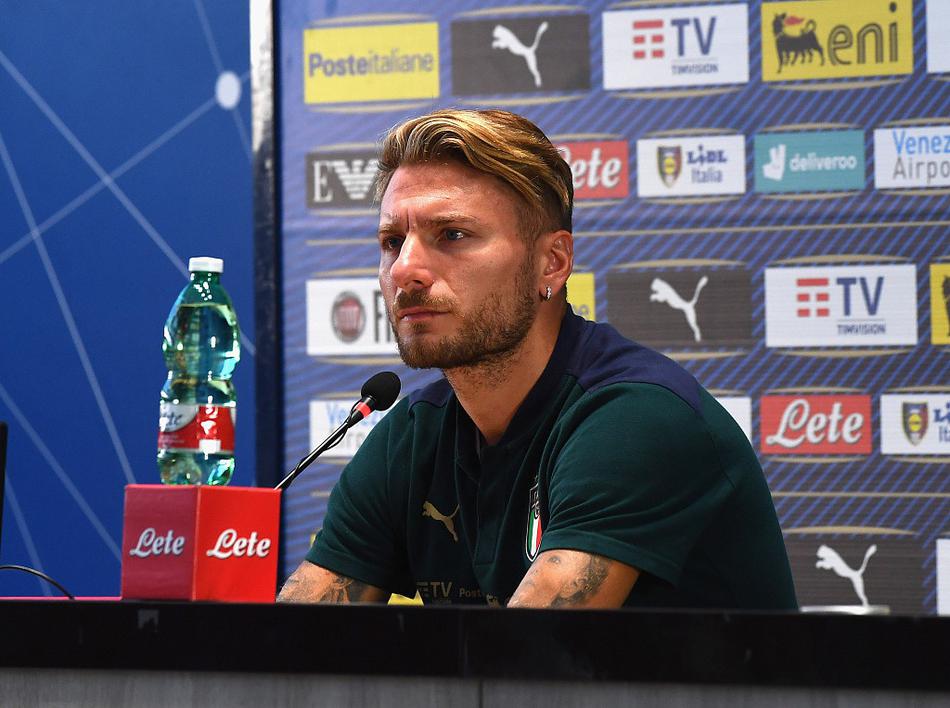 发布会现场的另一瞬间，因莫比莱侧身聆听记者提问，神情专注而放松，右手轻轻搭在桌面的麦克风上，桌上摆放着意大利国家队的官方水瓶，整个画面透露出大赛前宁静而专注的氛围。