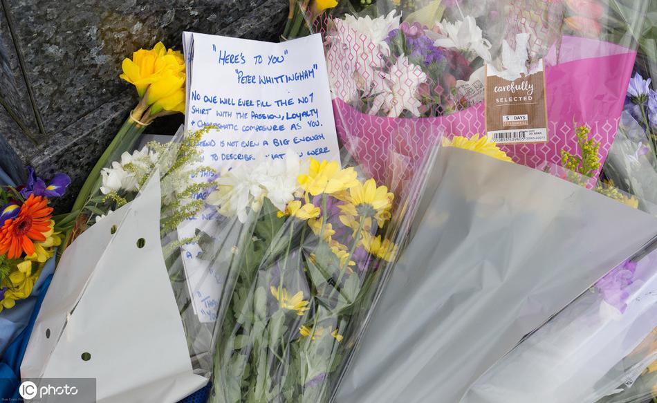 一位头发花白的老球迷，身穿厚厚的羽绒服，深情抚摸着惠廷厄姆雕像的底座，底座上摆满了球迷自发献上的蓝色围巾和鲜花，远处的加的夫城球场在阴天下显得庄严肃穆。