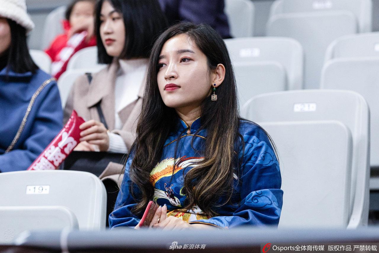 一位戴着眼镜的知性美女球迷，身着深色外套，在赛场灯光下静静观看比赛，她身前是热情澎湃的观众席，身后是激烈的赛场，动静之间尽显球迷文化的多元。