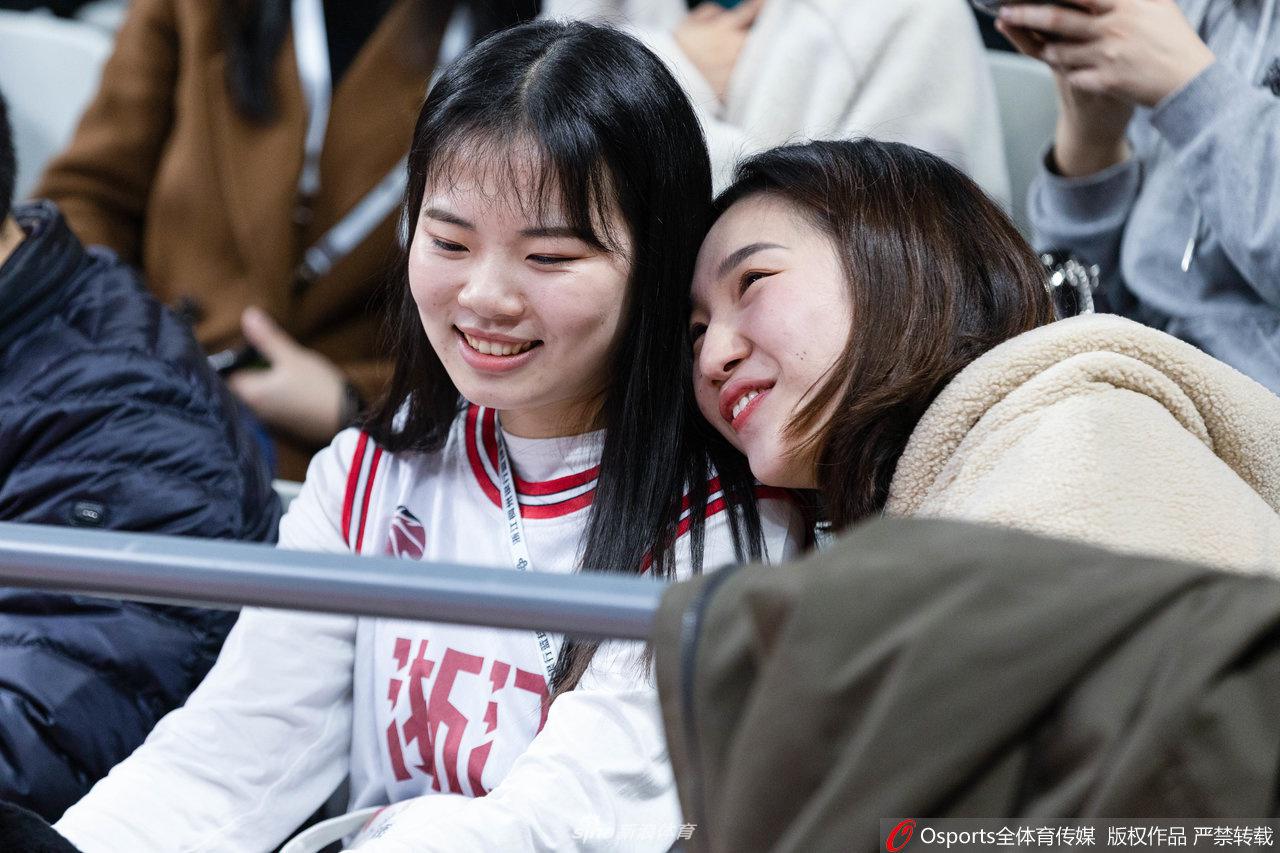 一位侧脸线条非常优美的新疆籍美女球迷，戴着精致的耳饰，目光越过人群投向远方，她的神情中带着一丝对球队暂时领先的欣慰，背景是虚化的赛场灯光。