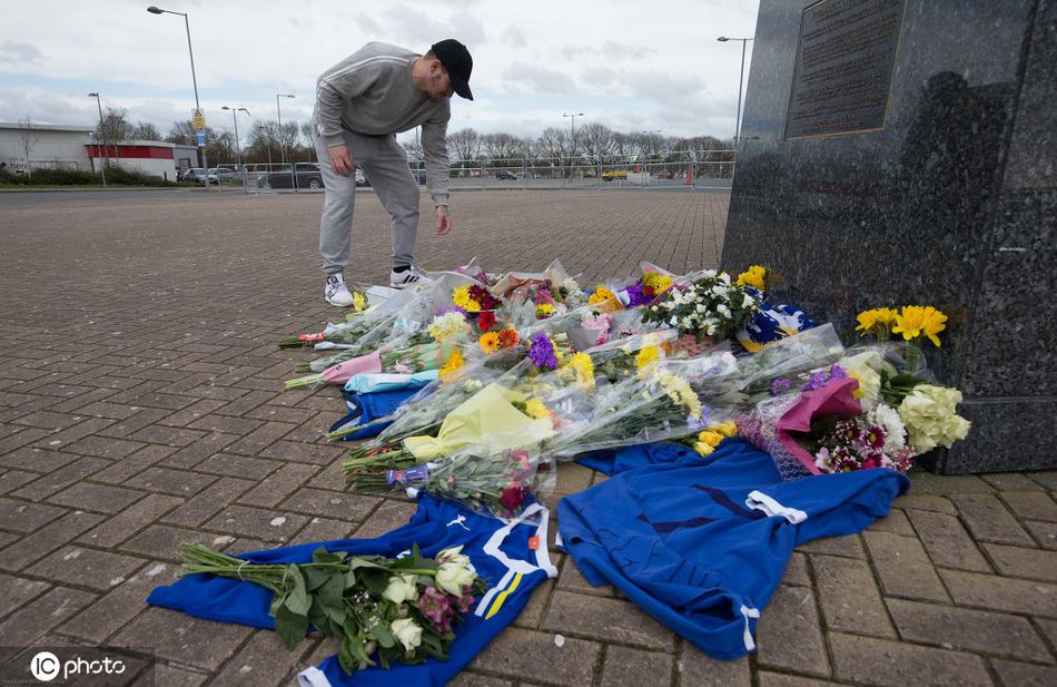 球迷将一件精致的惠廷厄姆7号球衣整齐地叠放在雕像脚下，旁边是一束含苞待放的白色百合花，球衣上的名字在闪光灯下折射出球迷无尽的思念。