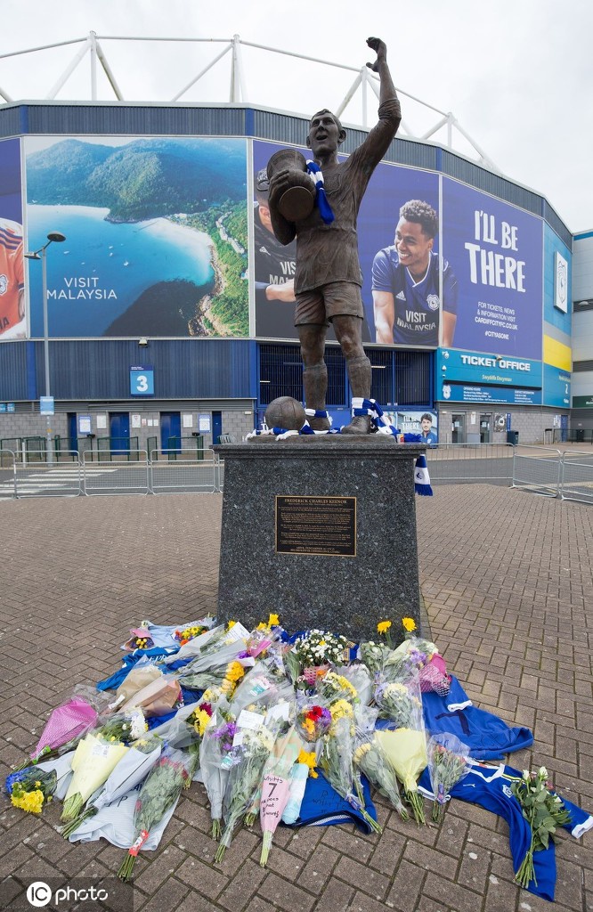 从雕像仰望的角度向上拍摄，天空中乌云开始散开，一缕阳光穿透云层洒在惠廷厄姆雕像的肩膀上，仿佛是天国投射下的圣光，温柔地拥抱这位早逝的足球天才。