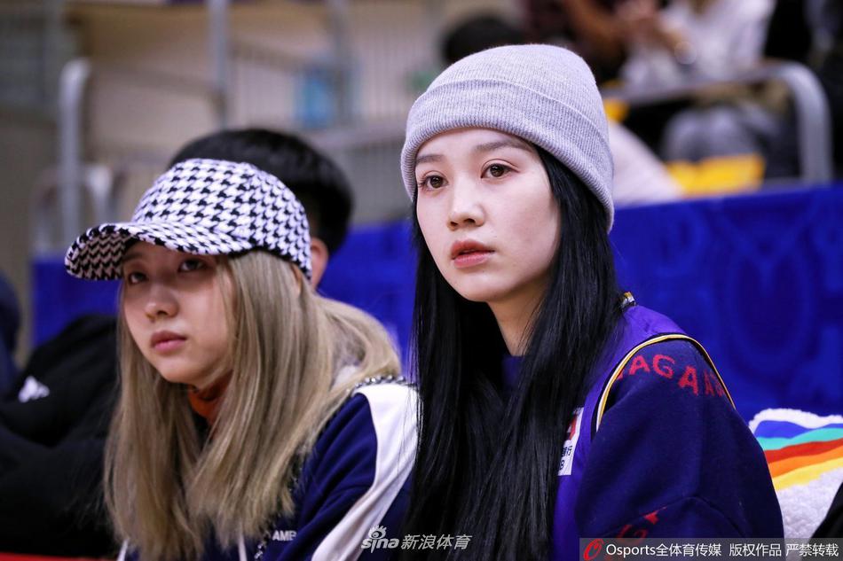 美女球迷手拿充气加油棒，脸上洋溢着灿烂的微笑，她身后是坐满了观众的看台，整个场馆内充满了热烈的比赛氛围。