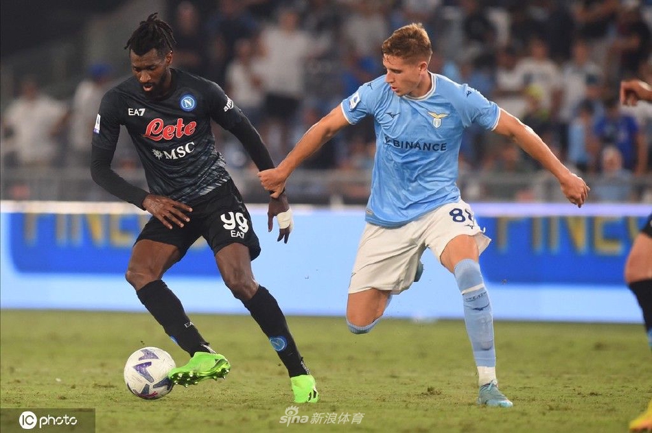 克瓦拉茨赫利亚进球后怒吼庆祝，身后是沮丧的拉齐奥后卫，背景看台上天蓝色旗帜舞动