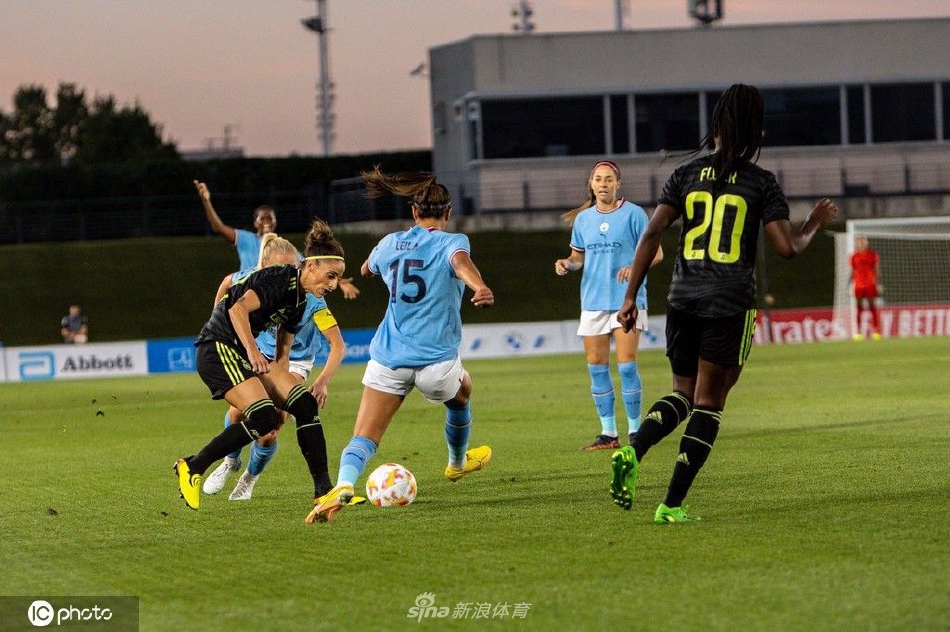 曼城女足球员在比赛末段带球突破，面对两名防守球员的包夹依然坚决内切，她的表情坚毅，步伐坚定，体现了永不放弃的体育精神。