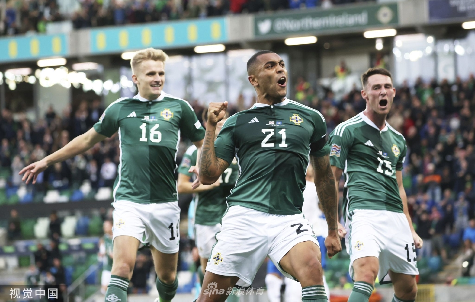 温莎公园球场全景，北爱尔兰与科索沃球员在绿茵场上激烈对抗，看台上坐满了挥舞旗帜的主队球迷。