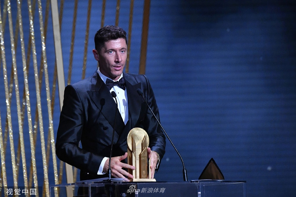 莱万手持奖杯，与一位身穿红色长裙的女士在典雅的剧院走廊中交谈。两人笑容温暖，气氛轻松愉快，展现了荣耀时刻之外的人际温情。