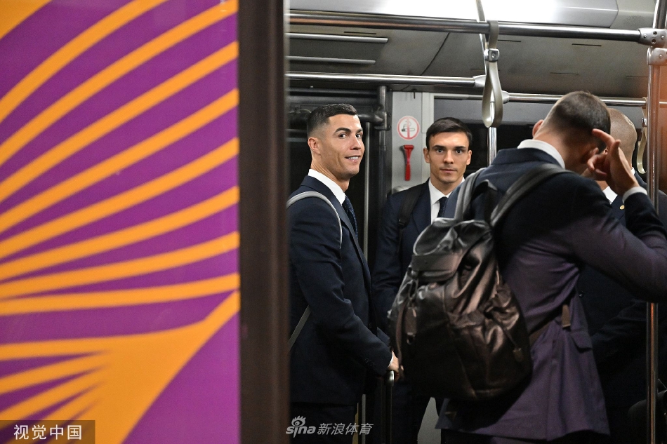 C罗与队友并肩走向接驳大巴，他侧头与身旁人交谈，嘴角带着一丝自信的微笑，手中拖着行李箱步履从容