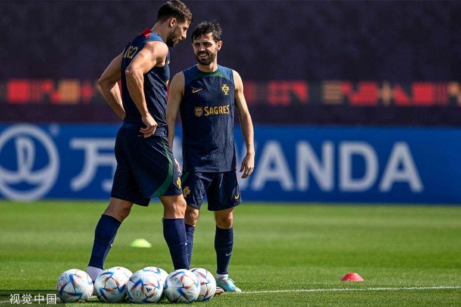 莱奥低头查看自己的球衣，A席的手停留在他肩头，背景是其他正在进行热身活动的葡萄牙队员，展现团队忙碌而有序的备战场景