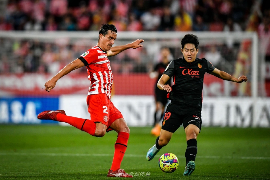 双方球员在中场激烈拼抢，赫罗纳球员正用身体护住皮球，马洛卡球员从侧后方伸脚试图断球，草屑在对抗中飞扬