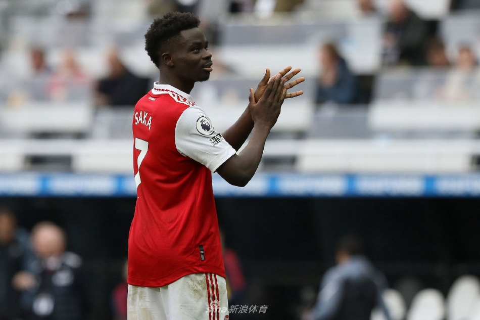 布卡约·萨卡手持阿森纳球衣，面带坚定而自豪的笑容，在签约桌前展示印有自己名字和2027年的新合约