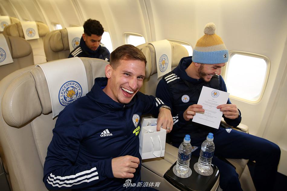 麦迪逊低头摆弄手机，试图在漫长的旅途中寻找一丝消遣，身后是队友模糊的身影与沉默的行李。