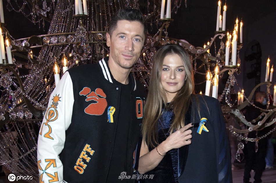 莱万与安娜在时装周入口处亲密相拥，莱万轻揽妻子腰肢，两人面向镜头露出幸福微笑，身后是古典的巴黎建筑背景。