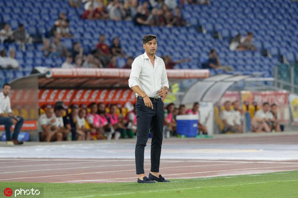 丰塞卡侧身面对球场，一只手举起指向球员，仿佛正在向场上队员下达最新的战术指令，背景是罗马奥林匹克球场坐满观众的看台。