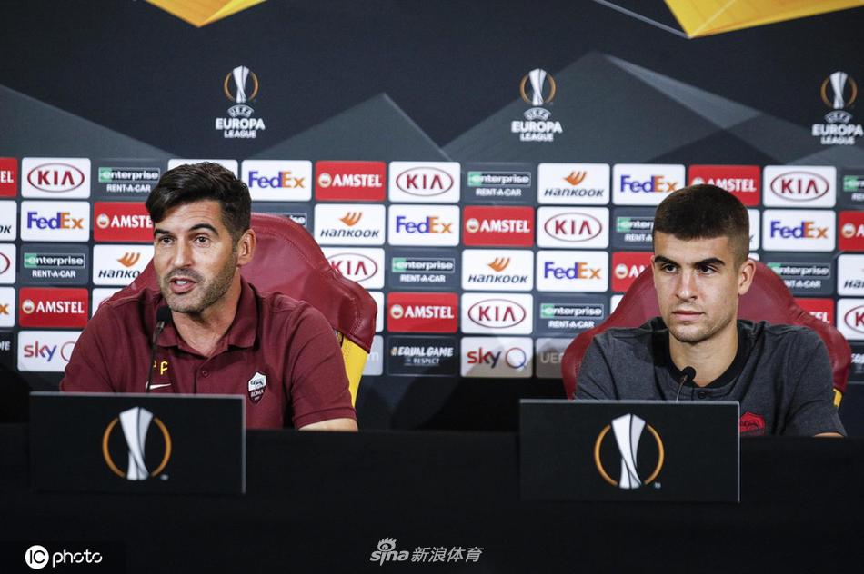 发布会现场全景，话筒林立，镜头聚焦，丰塞卡的身影在主席台中央显得沉稳而有力，背景的广告牌和队徽清晰可见，烘托出赛前紧张而正式的气氛。