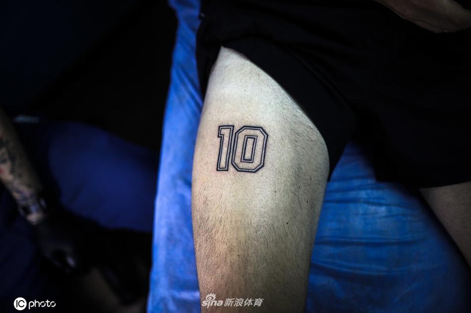 纹身店内灯光温暖，一位刚刚完成纹身的球迷低头凝视着自己小腿上崭新的“10号”图案，手指轻轻触摸，仿佛在感受偶像留下的最后温度，神情哀伤却充满慰藉。