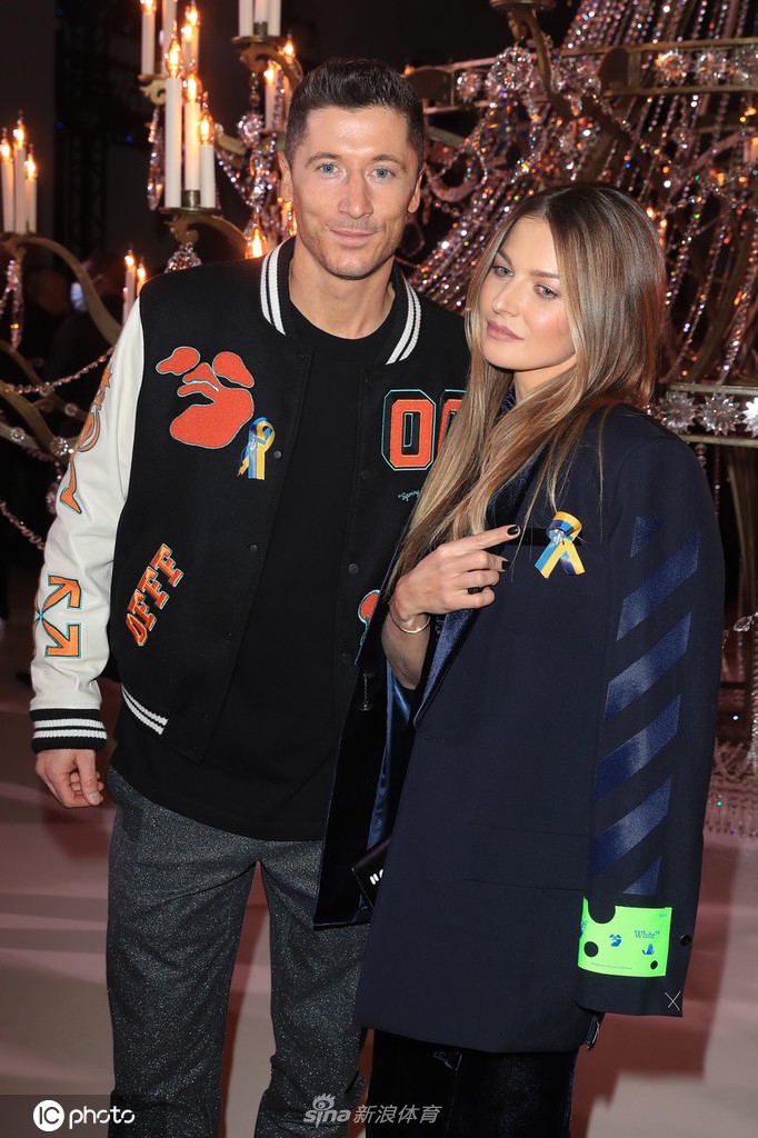 安娜挽着莱万的手臂走过石板路，风衣下摆随风轻扬，莱万侧身护着妻子，画面充满了动态的默契与保护感。