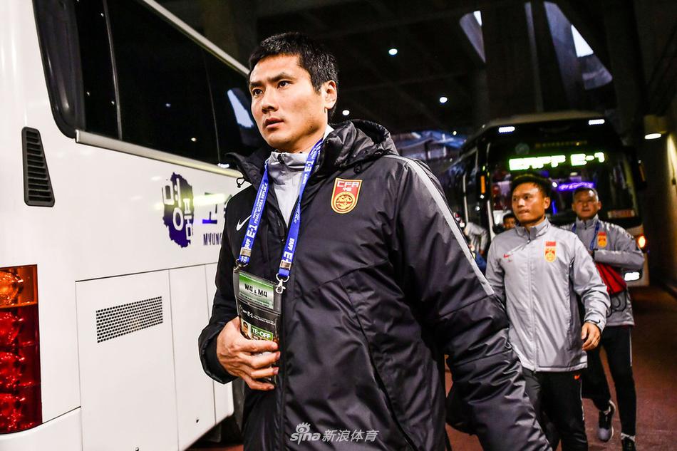 釜山寒夜中，身披红色战袍的国足选拔队将士们正围成一圈，聚精会神地聆听主教练李铁的战术部署，眼神中透露出对胜利的坚定渴望。