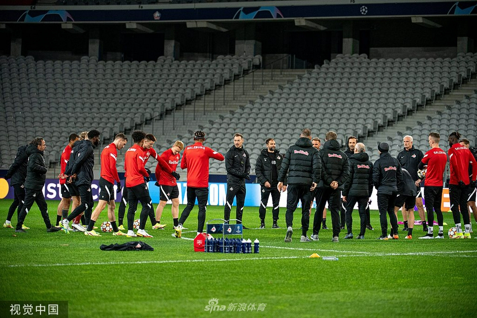 训练场边，一名萨尔茨堡球员正弯腰系鞋带，调整脚上的球鞋，额头上挂着细密的汗珠，阳光在他的球衣上反射出耀眼的红色光芒