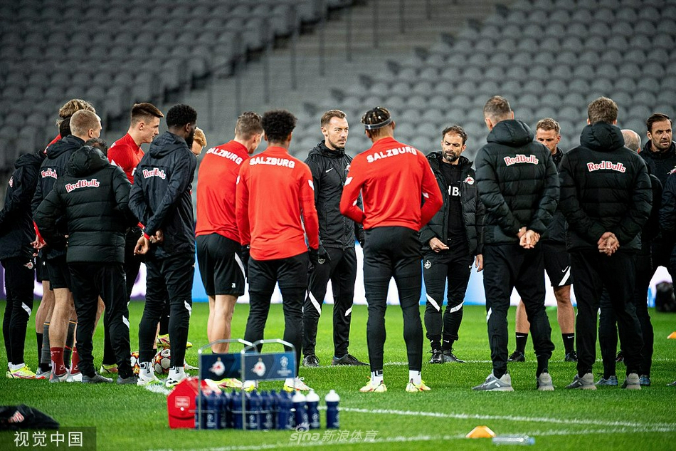 身穿白色教练组服装的主教练正在场边用手比划着战术，身后的球员们聚精会神地聆听，背景是法国冬日的低矮云层和看台的支架