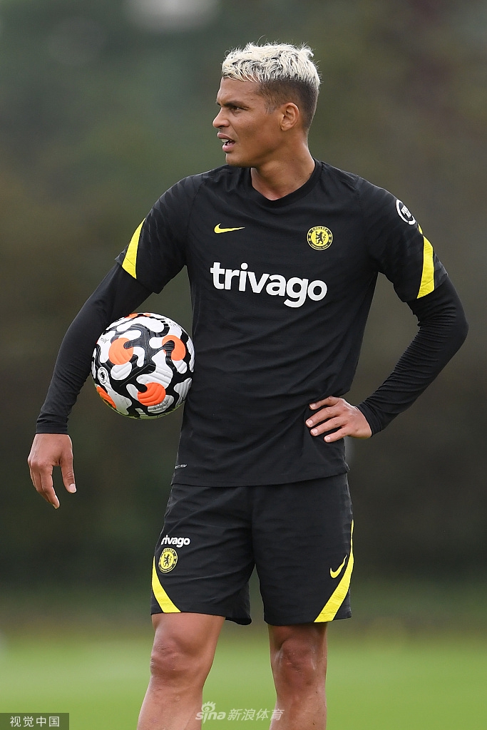 哈弗茨正低头专注地调整着自己的球袜，阳光勾勒出他坚毅的侧脸轮廓，背景中队友们模糊的身影和训练器材，更衬托出大战前夕的宁静与专注。