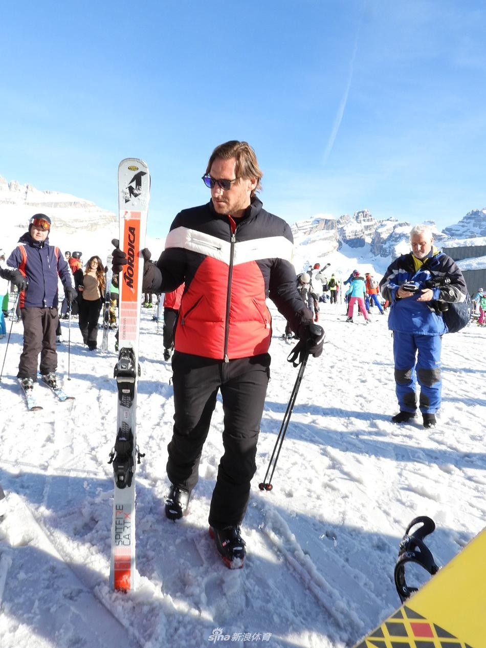 托蒂的小女儿坐在雪地上玩耍，手里捧着一团雪球，眼神纯真，托蒂则蹲在她身旁，温柔地为她整理雪镜。